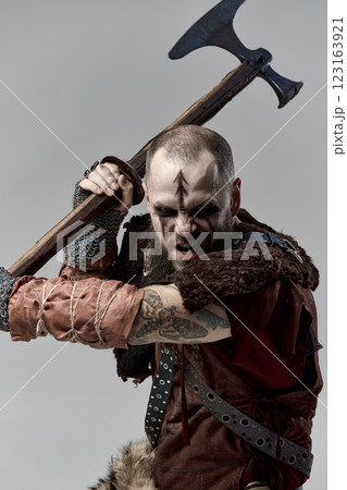 Medieval Viking warrior dressed in armor, gripping massive axe in both hands, poised to strike with fierce determination against studio background Medieval Viking warrior dressed in armor, gripping massive axe in both hands, poised to strike with fierce determination against studio background 123163921