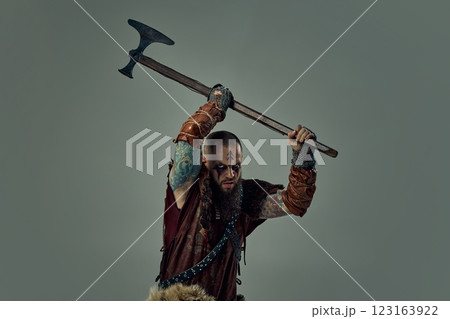 Fierce man, medieval Viking warrior in leather armor raising massive axe overhead, preparing for battle against studio background. Fierce man, medieval Viking warrior in leather armor raising massive axe overhead, preparing for battle against studio background. 123163922
