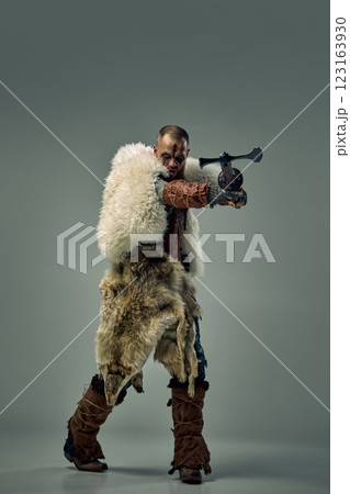 Fierce Viking warrior in medieval attire, in fur cloak, extending arm while holding sword aimed at enemy against studio background. Fierce Viking warrior in medieval attire, in fur cloak, extending arm while holding sword aimed at enemy against studio background. 123163930