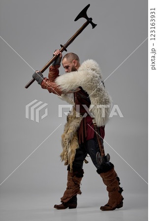 Medieval Viking warrior clad in fur cloak and armor, gripping massive axe in both hands, poised to strike with fierce determination against studio background. Medieval Viking warrior clad in fur cloak and armor, gripping massive axe in both hands, poised to strike with fierce determination against studio background. 123163931