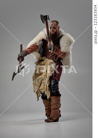 Warrior gripping two axes with intense gaze, man dressed in armor and fur cloak enhancing battle-ready stance against grey studio background. Warrior gripping two axes with intense gaze, man dressed in armor and fur cloak enhancing battle-ready stance against grey studio background. 123163934