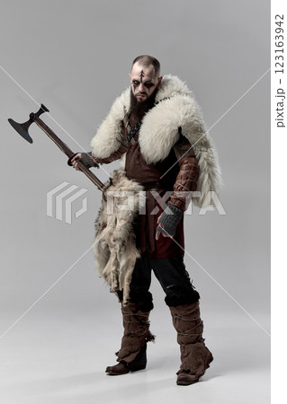 Full-length portrait of man, medieval Viking, warrior in armor, fur cloak, standing with axe against grey studio background Full-length portrait of man, medieval Viking, warrior in armor, fur cloak, standing with axe against grey studio background 123163942