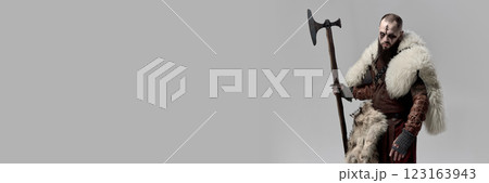 Portrait of medieval man, Viking in leather armor and fur cloak, standing with axe and gazing with determination against grey studio background. Portrait of medieval man, Viking in leather armor and fur cloak, standing with axe and gazing with determination against grey studio background. 123163943
