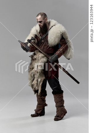 Full-length portrait of serious man, medieval Viking in leather armor and fur cloak, holding his axe against grey studio background. 123163944