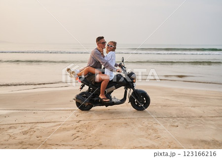 Couple on motorbike at beach. Man rides scooter with girlfriend hugging him. Sand, ocean waves in background. Honeymoon adventure travel concept. Romantic getaway on motorcycle by sea. Couple on motorbike at beach. Man rides scooter with girlfriend hugging him. Sand, ocean waves in background. Honeymoon adventure travel concept. Romantic getaway on motorcycle by sea. 123166216