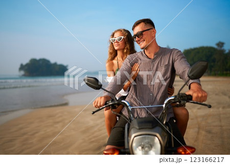 Smiling couple on motorbike at sunny beach. Enjoying ocean view, adventure travel. Man rides scooter. Romance, fun outdoors. Island escape, budget trip. 123166217