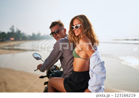 Smiling couple on motorbike at sunny beach. Young man and woman relax on motorcycle near ocean. Romantic adventure, love, travel by scooter, enjoy summer vibes, palm trees in background. 123166219