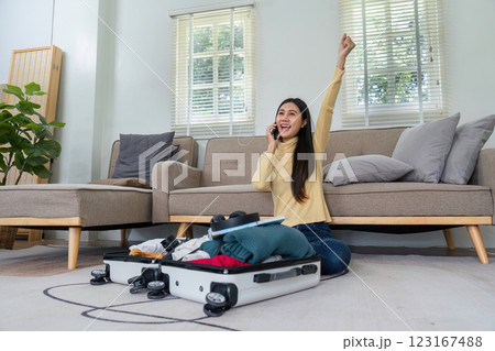Cheerful Young Woman Packing Suitcase for Travel While Talking on Phone in Modern Living Room Cheerful Young Woman Packing Suitcase for Travel While Talking on Phone in Modern Living Room 123167488