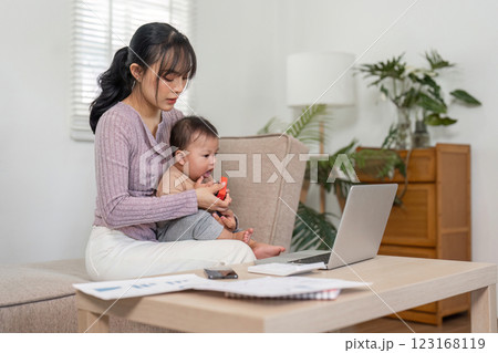 Mother working on laptop while holding baby and managing paperwork at home. 123168119