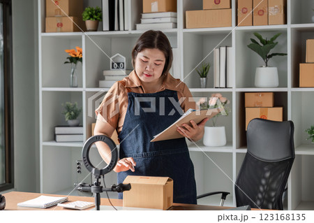 Young woman entrepreneur livestreaming to sell products online while reviewing orders in a stylish home office 123168135