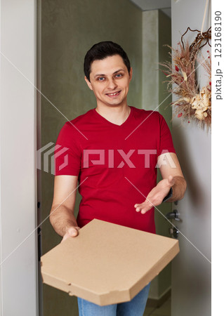 positive delivery man in red t-shirt giving food order, holding pizza box standing at doorway 123168190