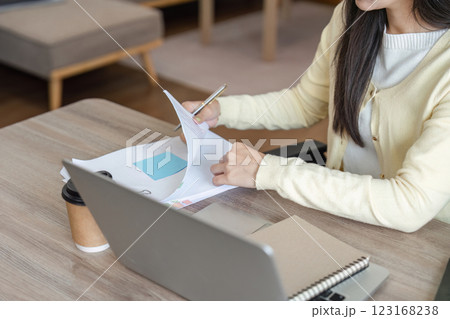 Focused Asian woman reviewing documents and taking notes while working on a laptop in a cozy home office. Focused Asian woman reviewing documents and taking notes while working on a laptop in a cozy home office. 123168238