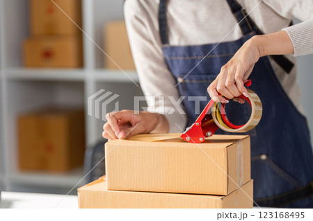 Close-up of young woman in apron sealing a shipping box with tape in a modern workspace. Close-up of young woman in apron sealing a shipping box with tape in a modern workspace. 123168495