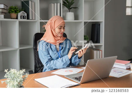Happy Muslim woman using smartphone while working on a laptop in an office. 123168583