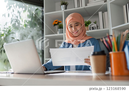 Smiling Muslim woman reviewing documents while working on a laptop in an office. Smiling Muslim woman reviewing documents while working on a laptop in an office. 123168590