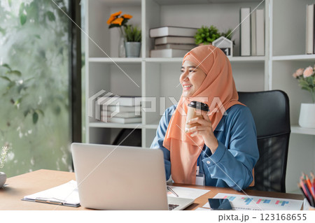 Muslim woman enjoying coffee while working on her laptop in a modern office. 123168655