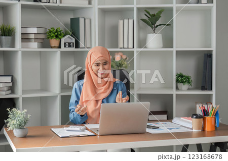 Muslim woman happily discussing in video call while working on a laptop at her desk. Muslim woman happily discussing in video call while working on a laptop at her desk. 123168678