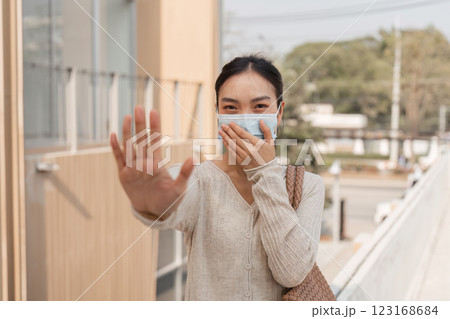 Diverse woman in a mask signaling stop while smiling outdoors in an urban setting. 123168684