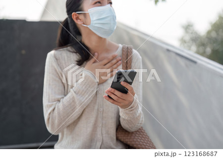 Young woman wearing a mask, holding smartphone, and showing emotion outdoors. 123168687