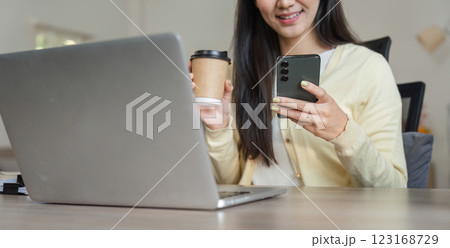 Happy Asian woman enjoying coffee while using smartphone and working on a laptop, reflecting a balanced work-life approach. Happy Asian woman enjoying coffee while using smartphone and working on a laptop, reflecting a balanced work-life approach. 123168729