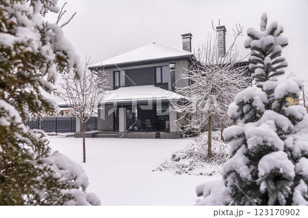 Modern two-story house with a snow-covered roof and patio surrounded by a winter garden with snow-laden trees and conifers. Modern two-story house with a snow-covered roof and patio surrounded by a winter garden with snow-laden trees and conifers. 123170902