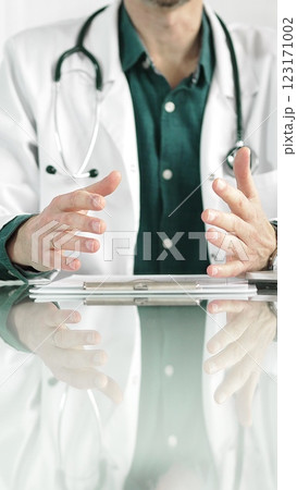 Doctor man wearing a stethoscope and lab coat is gesturing with hands while explaining a diagnosis to a patient across a glass desk with a laptop. Medicine concept Doctor man wearing a stethoscope and lab coat is gesturing with hands while explaining a diagnosis to a patient across a glass desk with a laptop. Medicine concept 123171002