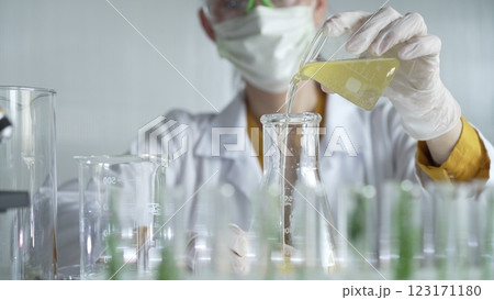 Female scientist researcher wearing a lab coat, white gloves, and mask, is pouring a yellow oily liquid from a beaker into an Erlenmeyer flask in laboratory, portrait. Science and medicine concepts 123171180