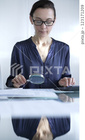 Woman accountant examining financial documents with magnifying glass at glass office desk, conducting thorough analysis of business transactions. Audit and taxes in business Woman accountant examining financial documents with magnifying glass at glass office desk, conducting thorough analysis of business transactions. Audit and taxes in business 123171209