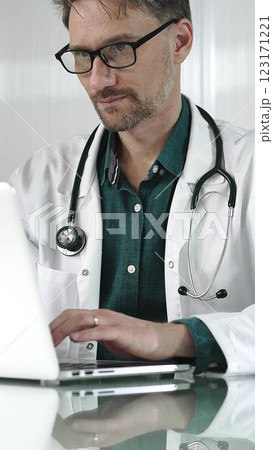 A male doctor in a green shirt under a medical coat is using technology in healthcare. Man is typing on a laptop on a glass desk in the clinic, vertical portrait view. Medicine A male doctor in a green shirt under a medical coat is using technology in healthcare. Man is typing on a laptop on a glass desk in the clinic, vertical portrait view. Medicine 123171221