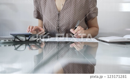 Businesswoman accountant, wearing brawn dotted blouse, is using a calculator to crunch numbers and jotting down meticulous notes, at sleek glass desk in a contemporary office. Taxes, audit in business 123171281