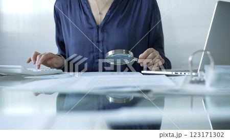 Businesswoman analyzing financial data with magnifying glass and calculator, performing accounting and auditing tasks in office. Audit and taxes theme in business 123171302
