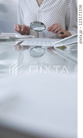 Businesswoman in white dotted blouse is carefully reviewing financial reports with magnifying glass and calculator, conducting audit and analyzing data, vertical view Taxes, audit in business 123171336