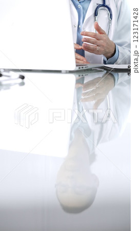 Unknown doctor woman, wearing a stethoscope, blue blouse under a lab coat, is gesturing with hands overs a glass desk with a laptop in clinic office, close up vertical view. Medicine and science 123171428