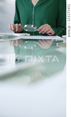 Businesswoman in green blouse carefully reviewing financial reports with magnifying glass and calculator, conducting audit and analyzing data in office. Taxes, audit in business Businesswoman in green blouse carefully reviewing financial reports with magnifying glass and calculator, conducting audit and analyzing data in office. Taxes, audit in business 123171451