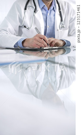 Unrecognizable doctor man in a blue shirt under a medical coat, is using a clipboard and taking notes on glass desk in a medical office, close-up vertical view. Medicine and health care Unrecognizable doctor man in a blue shirt under a medical coat, is using a clipboard and taking notes on glass desk in a medical office, close-up vertical view. Medicine and health care 123171461