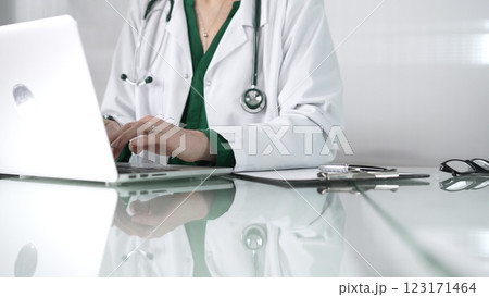 Female doctor using technology in healthcare, close up of hands typing on laptop with stethoscope, glasses, and clipboard on desk 123171464