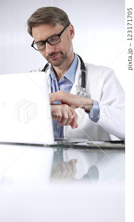 Doctor man in a blue shirt under a medical coat is thinking while using technology in medicine. Portrait of physician typing on a laptop on a glass desk in clinic, vertical view. Medicine and science Doctor man in a blue shirt under a medical coat is thinking while using technology in medicine. Portrait of physician typing on a laptop on a glass desk in clinic, vertical view. Medicine and science 123171705