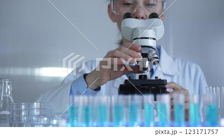 A female scientist, wearing a lab coat and safety glasses, is adjusting a binocular microscope surrounded by test tubes in laboratory setting. Medicine and science A female scientist, wearing a lab coat and safety glasses, is adjusting a binocular microscope surrounded by test tubes in laboratory setting. Medicine and science 123171757