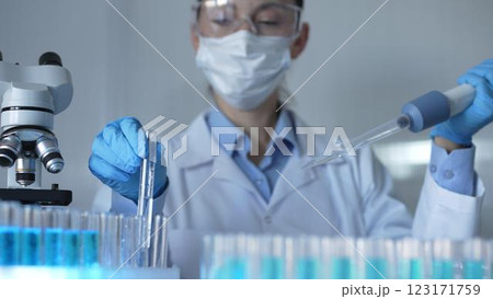 A female scientist, wearing a mask, blue gloves and safety glasses, is using a micropipette to drop a liquid into a test tubes. Medicine and science A female scientist, wearing a mask, blue gloves and safety glasses, is using a micropipette to drop a liquid into a test tubes. Medicine and science 123171759