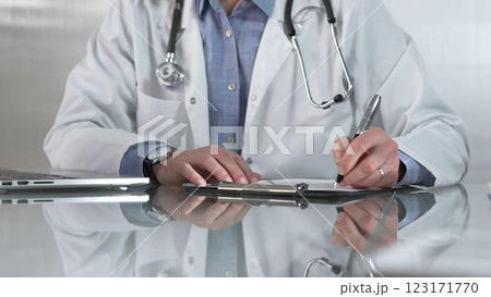 Male doctor, wearing blue shirt under a white medical coat, is using a clipboard and taking notes on glass desk in a medical office, close-up. Medicine concept Male doctor, wearing blue shirt under a white medical coat, is using a clipboard and taking notes on glass desk in a medical office, close-up. Medicine concept 123171770