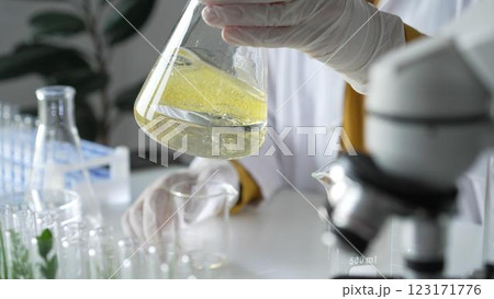 Unknown scientist wearing white protective gloves is carefully shaking up a yellow chemical solution inside an Erlenmeyer flask in a laboratory, close up. Science and medicine 123171776