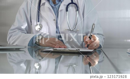 Woman doctor, wearing a blue blouse under a white medical coat, is using a clipboard and taking notes on glass desk in a medical office, close-up. Medicine 123171928