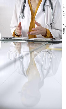 Unknown doctor woman, wearing a stethoscope and yellow blouse under a lab coat, is gesturing with hands overs a glass desk with a laptop in clinic office. Medicine and health care 123171936