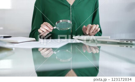 Businesswoman in green blouse carefully reviewing financial reports with magnifying glass and calculator, conducting audit and analyzing data in office. Taxes, audit in business 123172034