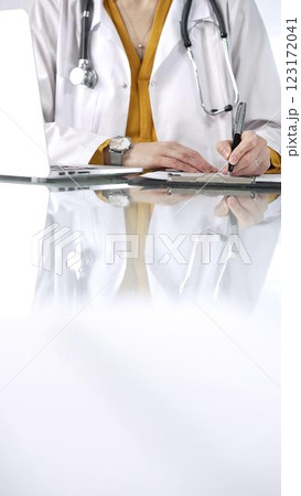 Unknown doctor woman, wearing a stethoscope and yellow blouse under a lab coat, is using a clipboard and taking notes on glass desk in a medical office, close-up vertical view. Medicine concept 123172041