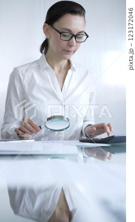 Businesswoman or accountant, wearing white casual shirt and glasses, is using magnifying glass and calculator to analyze financial documents in office setting. Audit and taxes in business 123172046