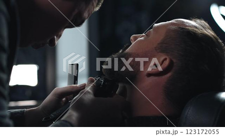 Skilled barber grooming client's beard using electric razor, delivering precise styling within contemporary barbershop setting 123172055