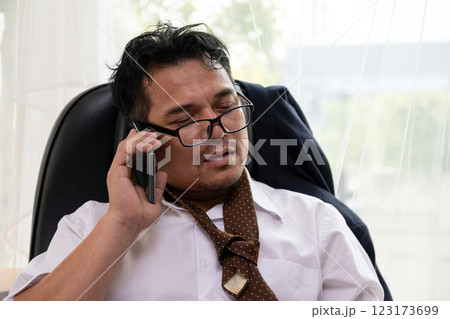 Exhausted businessman with messy appearance, loosened tie, and glasses askew talking on phone while sitting in office chair Exhausted businessman with messy appearance, loosened tie, and glasses askew talking on phone while sitting in office chair 123173699