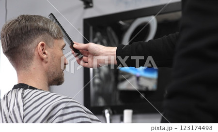 Barber carefully combing hair, client wearing protective cape, preparing for precision styling inside sleek contemporary barbershop setting 123174498