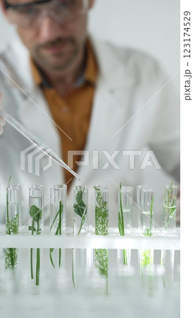 Man scientist wearing a lab coat, white gloves and protective glasses, is dripping liquid from a pipette into a test tube with a green plant inside, vertical close up view. Science and medicine 123174529